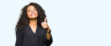 Kıvırcık saçlı zarif giyen genç güzel kız elbise hareketi el ile mutlu yaşasın yapıyor. Başarı gösteren kameraya bakarak ifade onaylama.