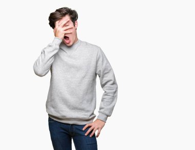 Mahcup ifadeyle parmaklarımın arasından seyir yüz ve gözler el ile kapsayan şok bakmak izole arka plan üzerinde sweatshirt giyen genç yakışıklı sportif adam.