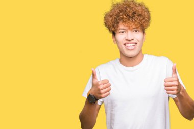El ile olumlu hareketi yaparken rahat beyaz t-shirt başarı işareti giyiyor afro saçlı yakışıklı delikanlı başparmak gülümseyen ve mutlu. Neşeli bir ifadeyle, kazanan jest kameraya bakıyor.