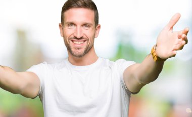 Yakışıklı adam sarılmak için kollarını açarak gülümseyerek kameraya bakarak casual beyaz t-shirt giyiyor. Neşeli ifade kucaklayan mutluluk.