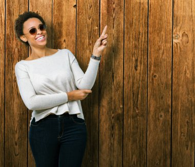 Kameraya bakarak yan el ve parmak ile işaret yüzünde kocaman bir gülümseme ile izole arka plan üzerinde güneş gözlüğü takıyor güzel genç Afro-Amerikan kadın.