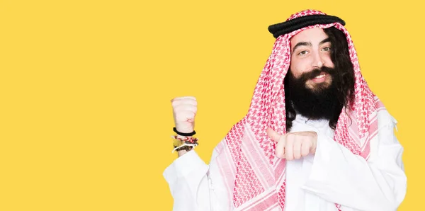 アラビア ビジネス伝統的なクーフィーヤを身に着けている長い髪の男は自信の笑みを浮かべて手と親指 裏側にポインティングをスカーフします — ストック写真