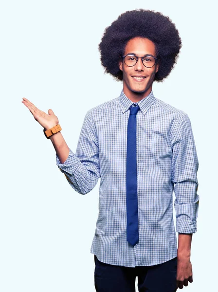 Young african american business man with afro hair wearing glasses smiling cheerful presenting and pointing with palm of hand looking at the camera.