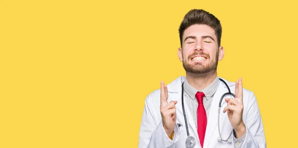 Joven Médico Guapo Con Abrigo Médico Sonriendo Cruzando Los Dedos —  Fotos de Stock