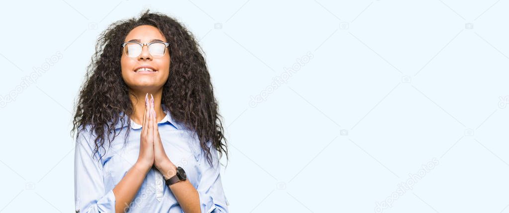Young beautiful business girl with curly hair wearing glasses praying with hands together asking for forgiveness smiling confident.