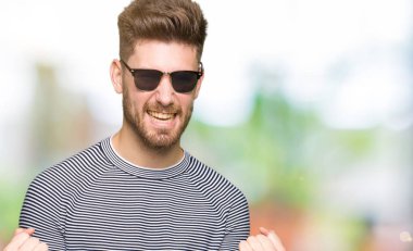 Gülümseyen ve başarı için çığlık çok mutlu ve heyecanlı kaldırdı, silah ile kazanan jest yapıyor güneş gözlüğü takıyor yakışıklı delikanlı. Kutlama kavramı.