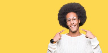 Afrikalı-Amerikalı genç afro saçlı gözlüklü emin gösterilen ve işaret parmakları diş ve ağız ile gülümseyen. Sağlık kavramı.