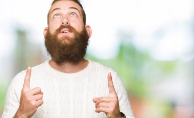 Kış kazak giyen genç hipster adam hayret ve yukarıya bakıyor ve parmakları ile işaret ve silah kaldırdı şaşırttı.