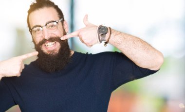 Uzun saç ve sakal emin gösterilen ve işaret parmakları diş ve ağız ile gülümseyen gözlüklü genç hipster adam. Sağlık kavramı.