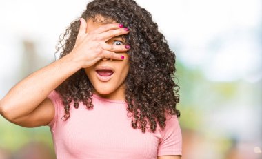 Pembe t-shirt giyen kıvırcık saçlı genç güzel kadın şok yüz ve gözleri el ile kaplayan gözetleme, utanmış ifade ile parmaklarından bakarak.