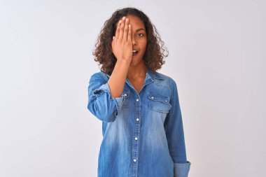 Genç Brezilyalı kadın kot gömlek giyiyor. İzole edilmiş beyaz arka planda duruyor. Bir gözünü eliyle kapatıyor, yüzünde kendine güvenen bir gülümseme ve sürpriz bir duygu var..