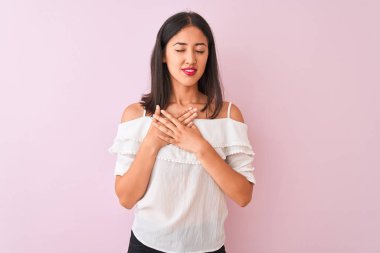 İzole edilmiş pembe arka plan üzerinde beyaz tişört giyen güzel Çinli kadın elleri göğsünde, gözleri kapalı ve yüzünde minnet dolu bir jest ile gülümsüyor. Sağlık kavramı.