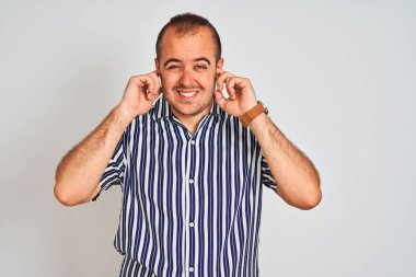 İzole edilmiş beyaz arka planda duran mavi çizgili tişört giyen genç adam yüksek sesli müzik sesi için kulakları parmaklarıyla kapatıyor. Sağır kavramı.