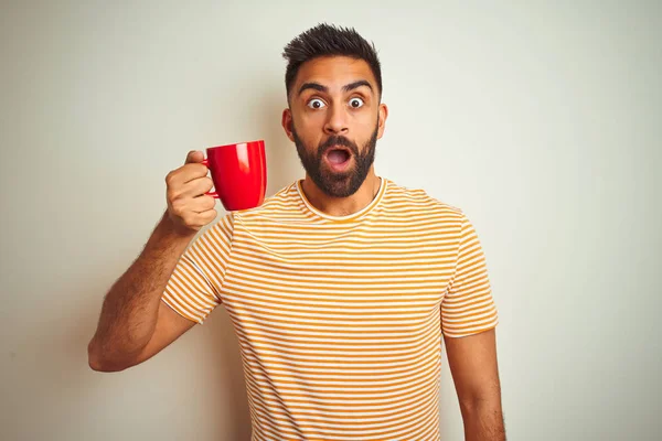 Young indian man drinking red cup of coffee standing over isolated white background scared in shock with a surprise face, afraid and excited with fear expression