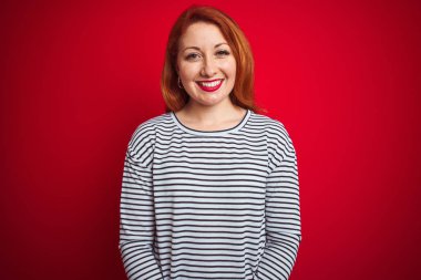 Kızıl saçlı genç bir kadın donanma gömleği giyiyor. Kırmızı izole edilmiş arka plan üzerinde duruyor. Yüzünde mutlu ve havalı bir gülümseme var. Şanslı kişi..