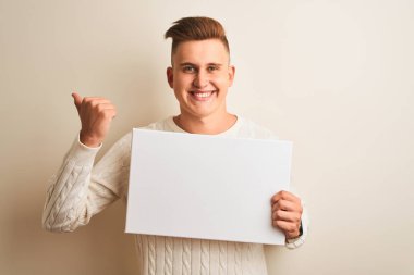 Genç yakışıklı adam izole beyaz arka plan üzerinde duran afiş tutan işaret ve mutlu yüzü gülümseyerek yan başparmak ile gösteren