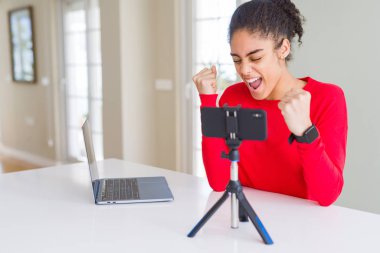 Akıllı telefon kamerasını kullanarak görüntülü görüşme yapan genç Afro-Amerikan kadın çok mutlu ve heyecanlı bir şekilde kollarını kaldırarak, gülümseyerek ve başarı için çığlık atarak kazanan jest yapıyor. Kutlama konsepti.