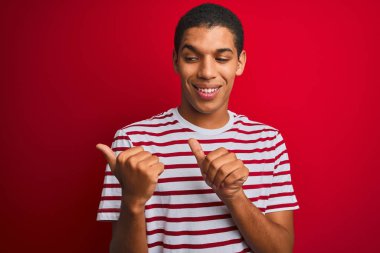 İzole edilmiş kırmızı arka plan üzerinde çizgili tişört giyen genç, yakışıklı bir Arap. Arka tarafı işaret ediyor. El ve başparmakları havada, kendinden emin.