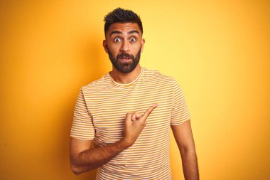 İzole sarı arka plan üzerinde duran t-shirt giyen genç hintli adam işaret parmağı ile endişeli ve sinirli bir kenara işaret, endişeli ve sürpriz ifade