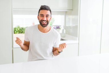 Evde yakışıklı İspanyol adam Casual beyaz t-shirt sürpriz kutluyor ve silah yükseltilmiş ve açık gözleri ile başarı için hayran. Kazanan kavramı.