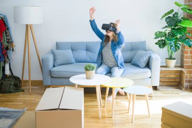 Yeni bir eve hareket karton kutular etrafında kanepede oturan bir simülasyon oyunu oynarken sanal gerçeklik gözlük giyen genç sarışın kadın