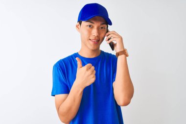 Büyük bir gülümseme ok işareti yapıyor mutlu izole beyaz arka plan üzerinde akıllı telefonda konuşurken kap giyen Çinli teslimatçı, parmakları ile başparmak, mükemmel işareti