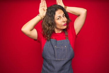 Orta yaş yaşlı kadın kırmızı izole arka plan üzerinde önlük üniforma giyen el avuç içi alaycı ve şüpheci görünümlü tavşan kulakları jest yapıyor. Paskalya tavşanı kavramı.