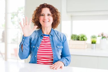Kıvırcık saçlı, kot ceket giyen orta yaşlı bir kadın beş numaralı parmaklarıyla işaret ederken kendinden emin ve mutlu gülümsüyor..