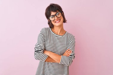İzole pembe arka plan üzerinde çizgili t-shirt ve gözlük giyen genç güzel kadın kameraya bakarak çapraz kolları ile gülümseyen mutlu yüz. Pozitif kişi.