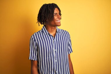Izole sarı arka plan üzerinde rahat çizgili t-shirt giyen dreadlocks ile Afro adam yüzünde gülümseme, doğal ifade ile yan bakıyor. Gülmekten emin.