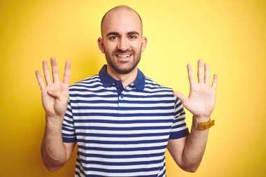 Sakallı genç kel adam sarı izole arka plan üzerinde rahat çizgili mavi t-shirt giyen gösteren ve emin ve mutlu gülümserken parmakları sayısı dokuz ile işaret.