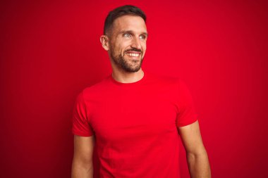 Genç yakışıklı adam kırmızı izole arka plan üzerinde rahat t-shirt giyen yüzünde gülümseme, doğal ifade ile yan bakıyor. Gülmekten emin.
