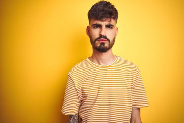 Çizgili t-shirt giyen dövme giyen genç adam izole sarı arka plan şüpheci ve sinir üzerinde duran, sorun nedeniyle üzgün kaşlarını çatma. Negatif kişi.