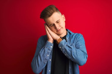 Denim gömlek giyen genç yakışıklı adam izole kırmızı arka plan üzerinde duran yorgun rüya ve kapalı gözlerle gülümseyerek birlikte elleri ile poz.