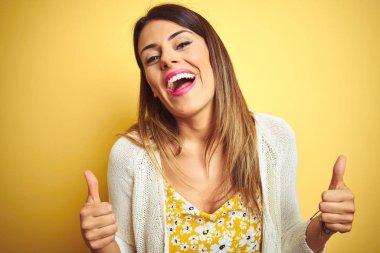 Sarı arka plan başarı işaretinin üzerinde duran ceketli genç ve güzel bir kadın. Elleriyle olumlu jest yapıyor. Gülümsüyor ve mutlu. Neşeli ifade ve kazanan jest.