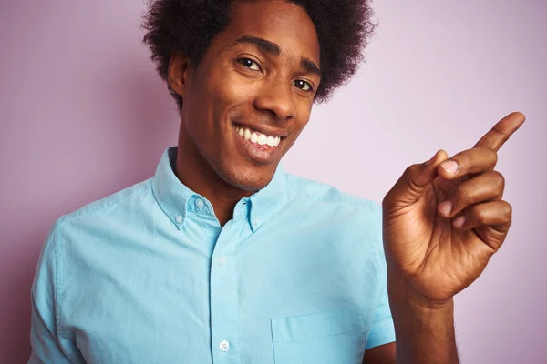 Young american man with afro hair wearing blue shirt standing over isolated pink background very happy pointing with hand and finger to the side