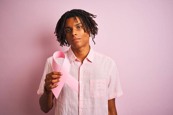 Afro american man with dreadlocks holding cancer ribbon over isolated pink background with a confident expression on smart face thinking serious