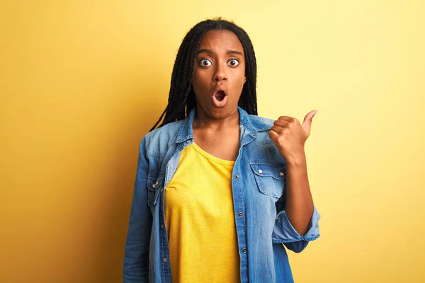 Young african american woman wearing denim shirt standing over isolated yellow background Surprised pointing with hand finger to the side, open mouth amazed expression.