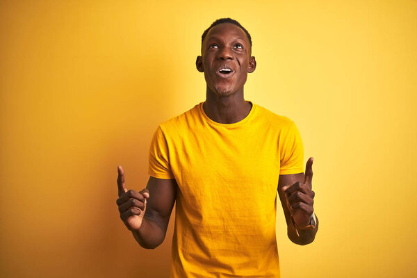 Young african american man wearing casual t-shirt standing over isolated yellow background amazed and surprised looking up and pointing with fingers and raised arms.