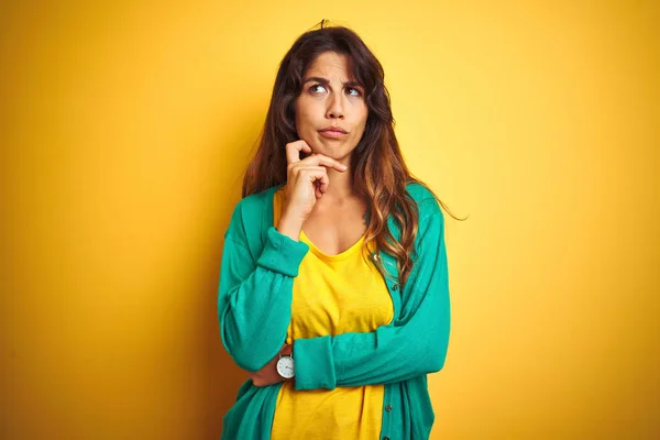 若い女性のTシャツと緑のセーターを着て 黄色の孤立した背景の上に立って質問については 集中的な表現を考えて顎の手で 思慮深い顔で微笑んだ 疑いの概念 — ストック写真