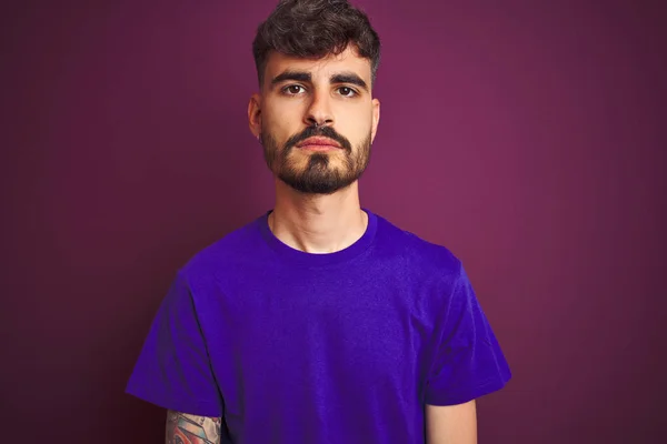 孤立した紫色の背景の上に立つタトゥーを着た若い男は 顔に真剣な表情でリラックスしました カメラを見てシンプルで自然な — ストック写真