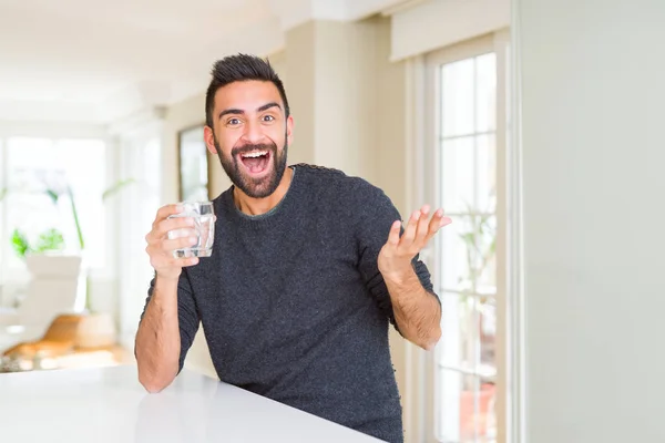 水の新鮮なグラスを飲むハンサムなヒスパニックの男は非常に幸せで興奮し 大きな笑顔で叫んで勝利を祝う勝者の表情と手を上げた — ストック写真