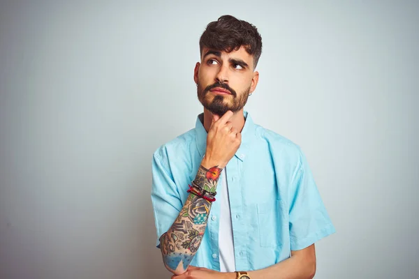 Young man with tattoo wearing blue shirt standing over isolated white background with hand on chin thinking about question, pensive expression. Smiling with thoughtful face. Doubt concept.