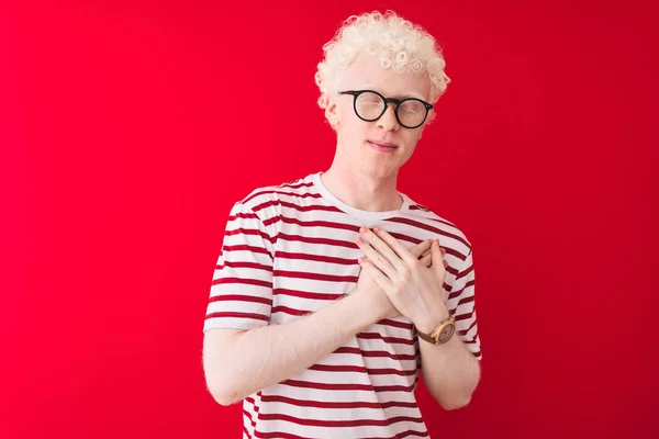Young albino blond man wearing striped t-shirt and glasses over isolated red background smiling with hands on chest with closed eyes and grateful gesture on face. Health concept.