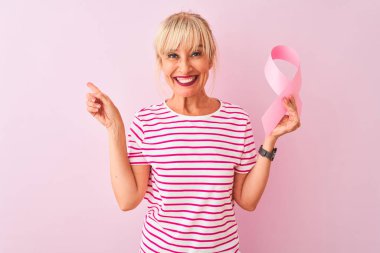Orta yaş kadın izole pembe arka plan üzerinde duran kanser kurdele tutan çok mutlu yan el ve parmak ile işaret