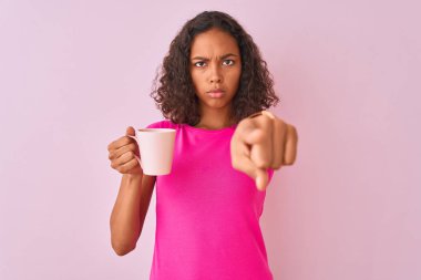 Genç Brezilyalı bir kadın izole edilmiş pembe arka planda durup kahve içerken kamerayı işaret ediyor ve sen, el işareti, ön taraftan pozitif ve kendinden emin bir hareket.