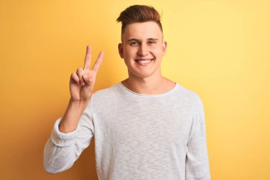 İzole edilmiş sarı arka plan üzerinde duran beyaz tişörtlü genç ve yakışıklı bir adam iki numarayı gösterirken kendinden emin ve mutlu bir şekilde gülümsüyor..
