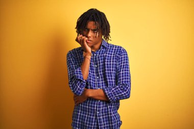 Yalnız sarı arka plan üzerinde duran rahat gömlek giyen dreadlocks ile Afro adam stresli ve ağız tırnak ısırma elleri ile sinir görünümlü. Anksiyete sorunu.