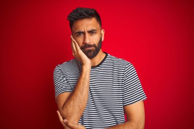 Genç yakışıklı hintli adam izole kırmızı arka plan üzerinde lacivert çizgili t-shirt giyen yorgun görünümlü ve çapraz kolları ile depresyon sorunları ile sıkılmış düşünme.