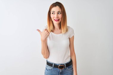 Güzel kızıl saçlı kadın izole beyaz arka plan üzerinde duran rahat t-shirt giyen mutlu yüz görünümlü ve başparmak ile yan işaret gülümseyerek.